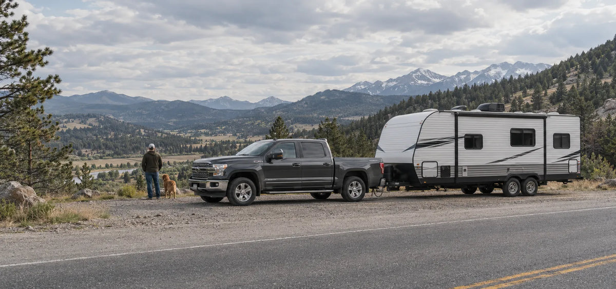 A dark gray pickup truck towing a white travel trailer parked on a roadside with mountains and pine trees in the background. A person and dog stand near the truck on the left side of the road.