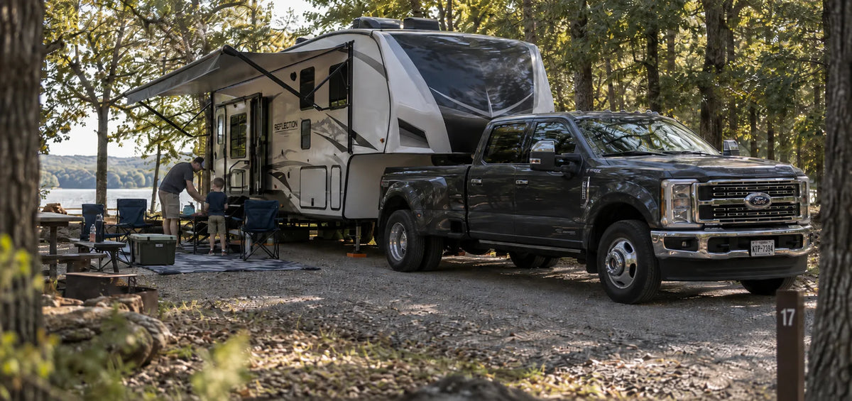 A black Ford pickup truck parked at a lakeside campsite next to a large white and gray travel trailer, with a person standing by the trailer entrance and trees surrounding the scene.